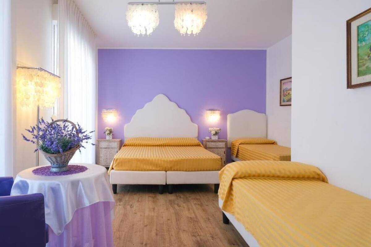 Ein Hotelzimmer mit violettem Wandhintergrund, zwei Betten mit gelber Bettwäsche, einem runden Tisch mit violetter Tischdecke und einer Blumenvase.