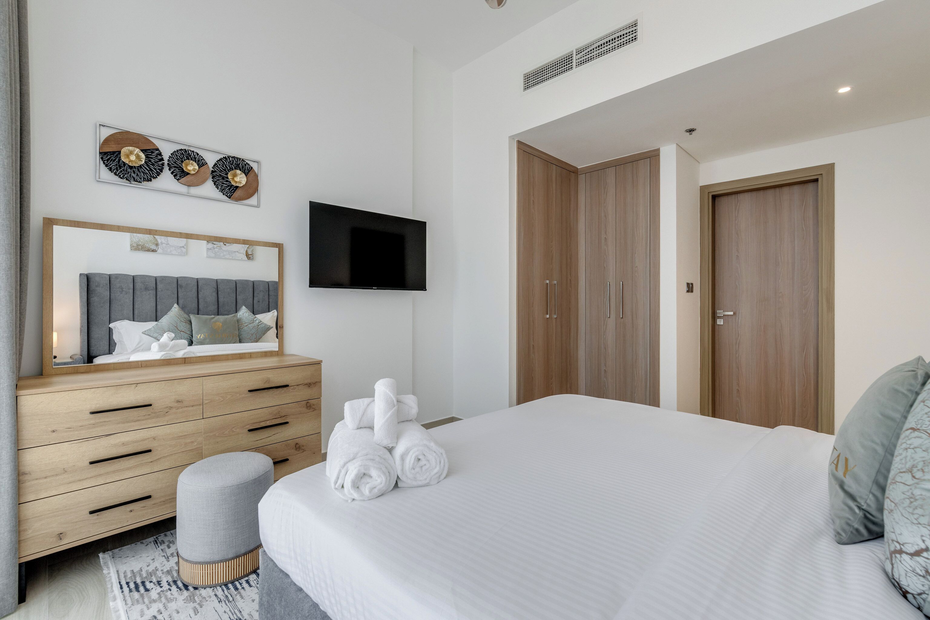 A modern bedroom with a large bed, wooden wardrobe, a flat-screen TV, and a mirror above the dresser.