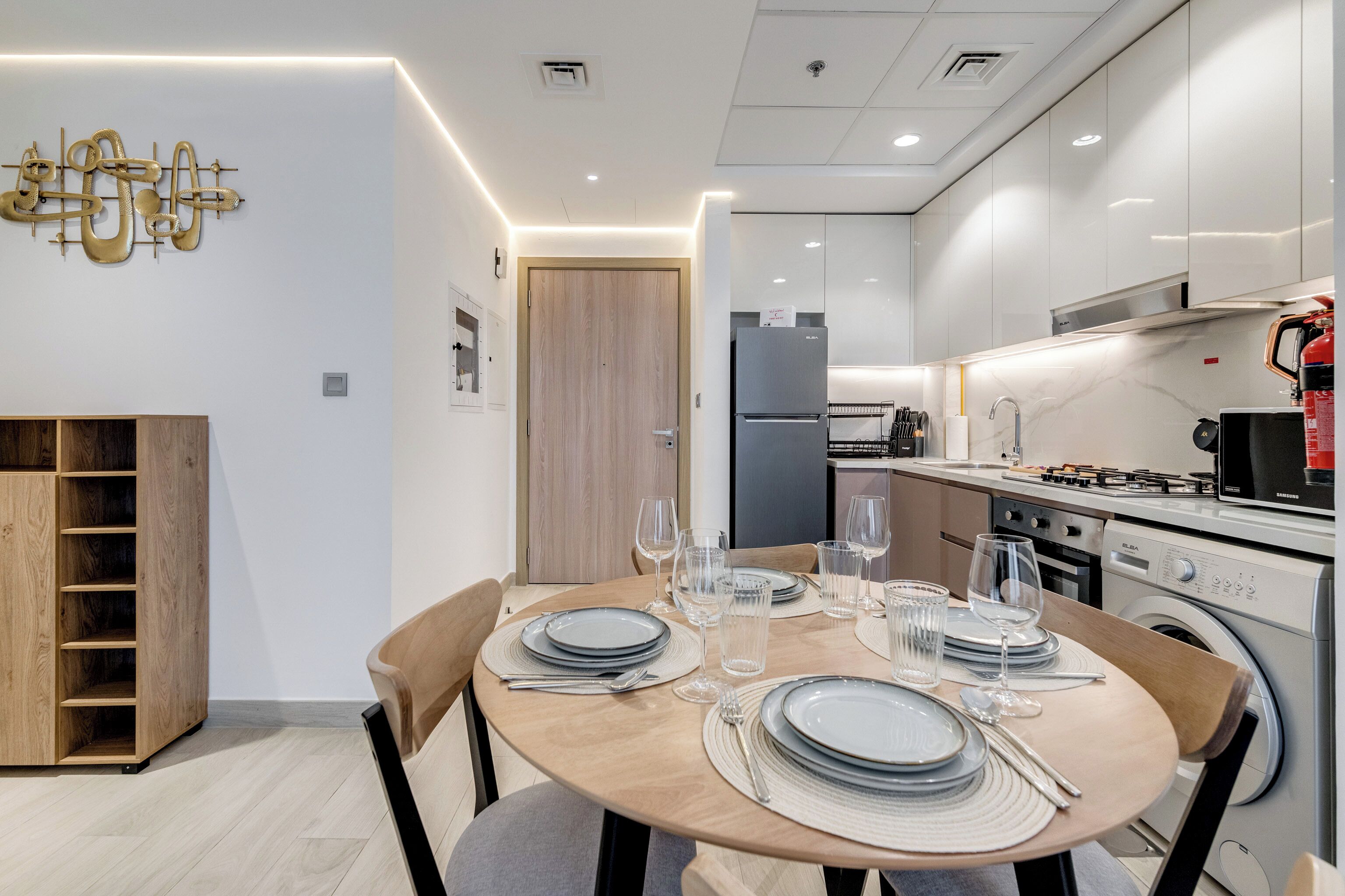 Una cocina moderna con armarios blancos, horno empotrado y microondas. Cuenta con un comedor con mesa redonda dispuesta para cuatro personas, incluyendo platos, vasos y cubiertos.