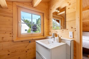 Interior de una cabaña de madera con baño que incluye ventana, lavamanos, espejo y cama.