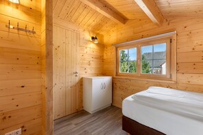 Una habitación con paneles de madera, una cama, un mueble blanco pequeño, una ventana y una lámpara fijada a la pared.