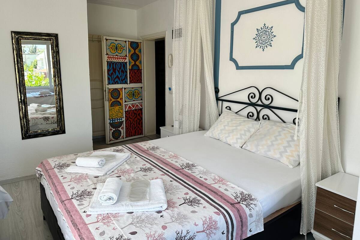 A neatly made bed with a patterned bedspread and pillows, a bedside table, a mirror, and a door with colorful tiles.
