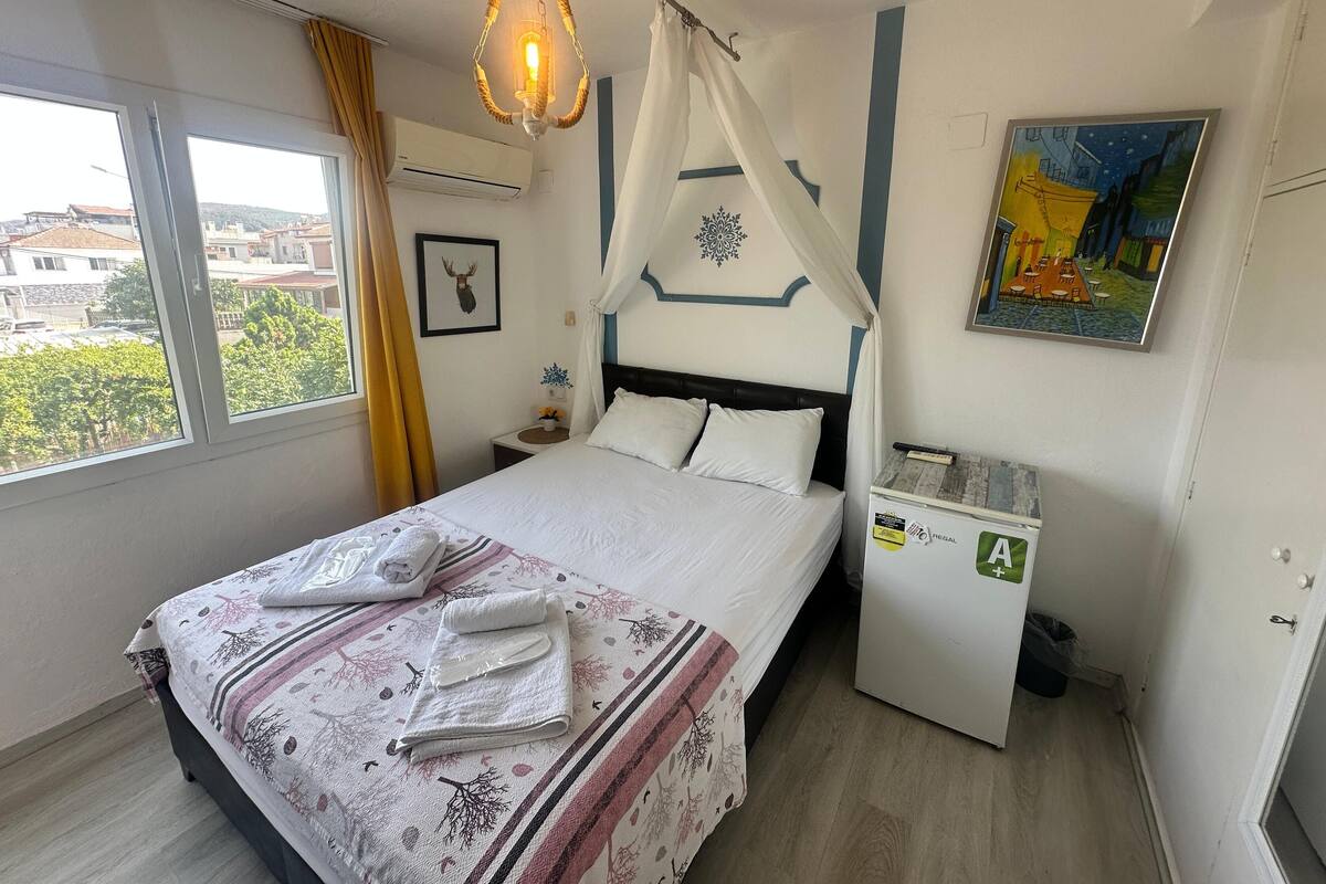 A neatly made bed with white linens and a patterned blanket, a bedside table with a lamp, a wall-mounted air conditioner, and a window with curtains.