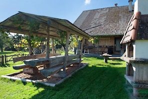 Un espace extérieur aménagé dans un style rustique, avec un banc en bois et une table de pique-nique couverte, une clôture en bois et un bâtiment doté d’un toit en tuiles.