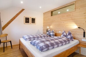 A bedroom with a wooden headboard, two beds with checkered bedding, a wooden chair, and a bedside table with a lamp.