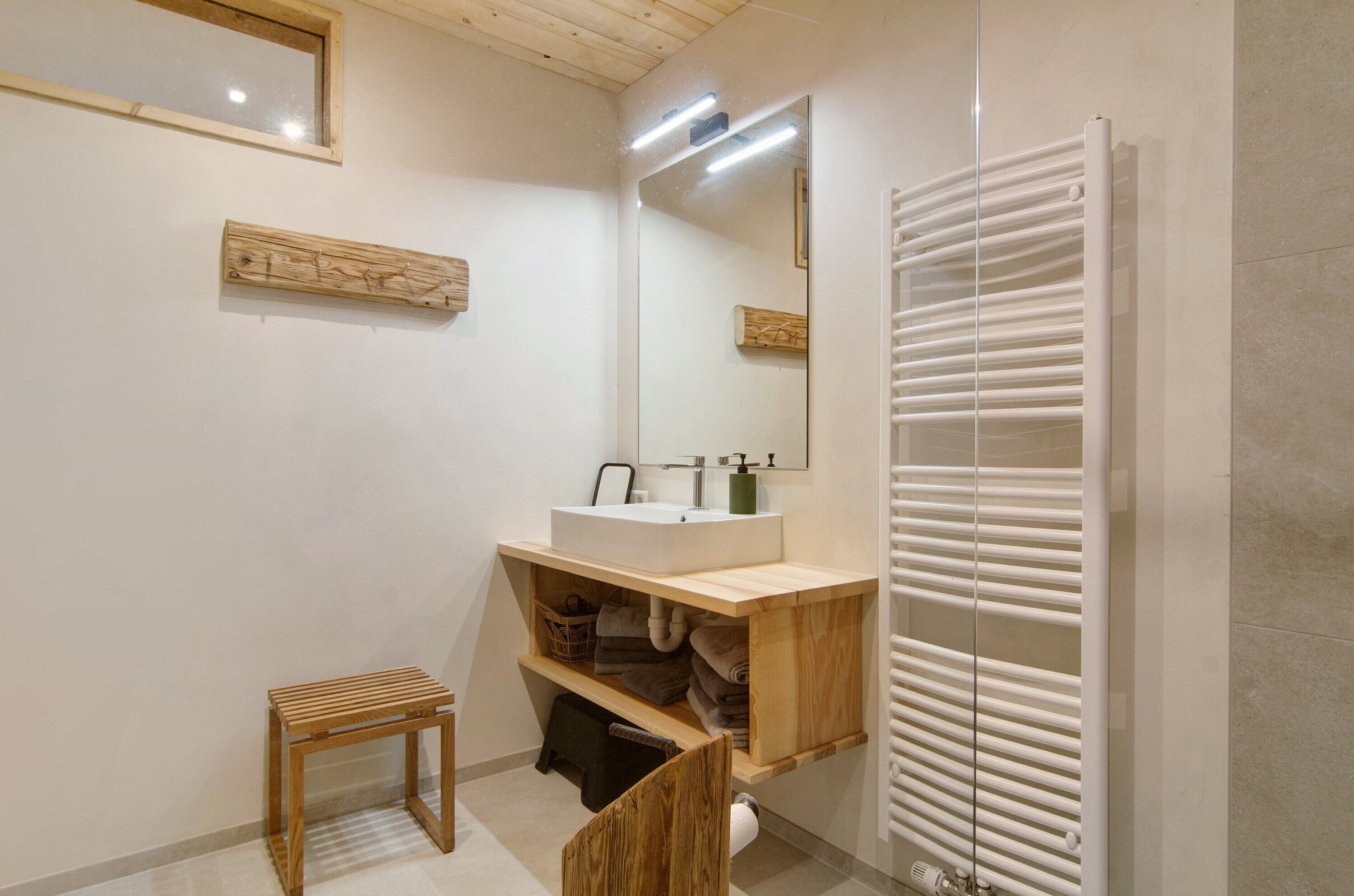 Een badkamer met een houten wastafelmeubel, een grote spiegel, een handdoekrek en een klein houten krukje.