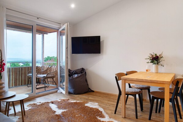 Une pièce comprenant une table à manger en bois, des chaises, un téléviseur fixé au mur, une grande fenêtre offrant une vue dégagée et une porte-fenêtre coulissante donnant sur un espace extérieur aménagé avec des meubles en osier.