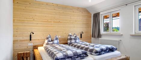 A room with a wooden slat wall, two beds with checkered bedding, a bedside table, a lamp, and a window with curtains.