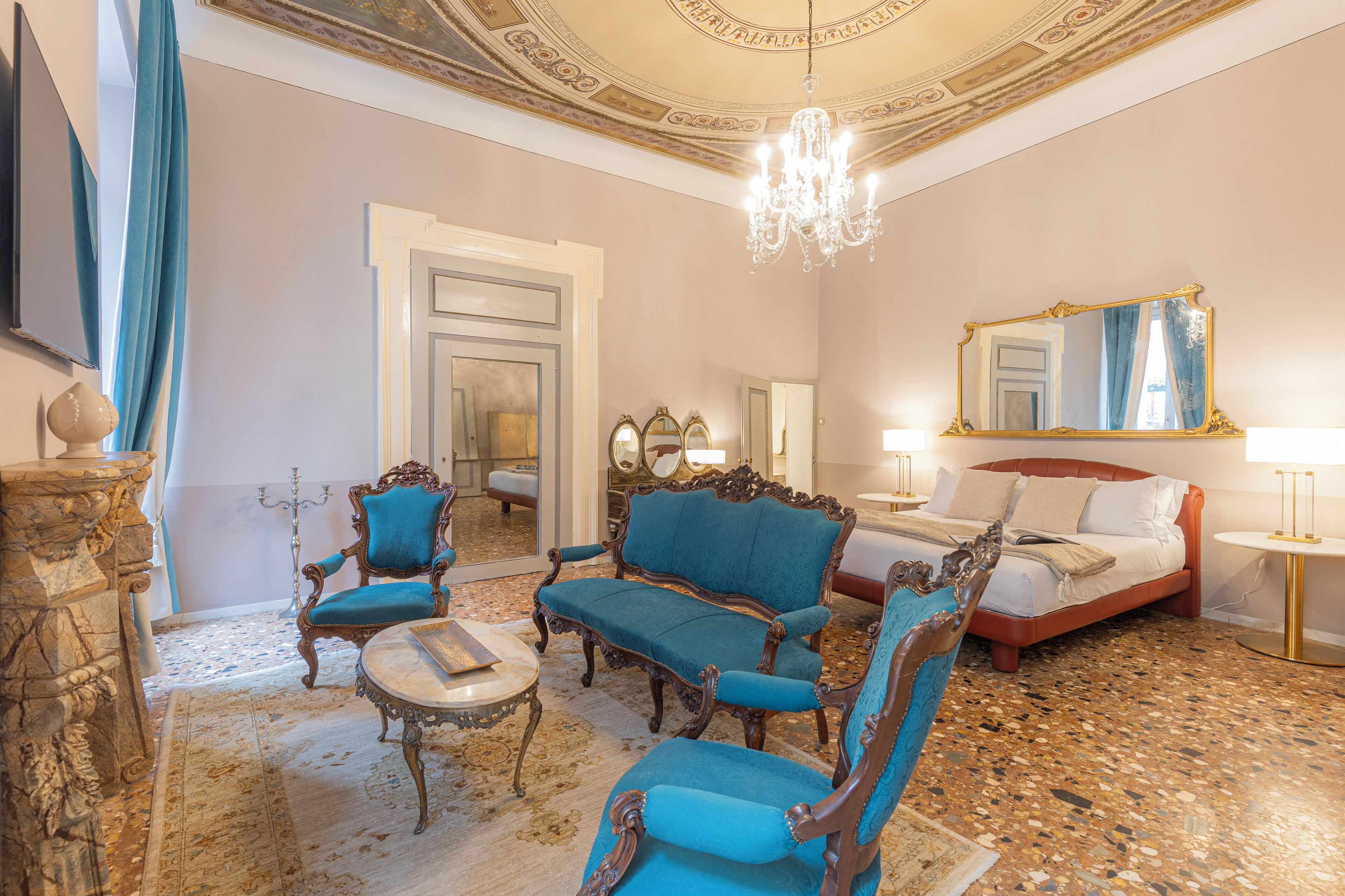 A room with a bed, two blue armchairs, a round table, and a chandelier.