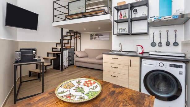Een ruimtelijk ingerichte leefruimte met een hoogslaper, kitchenette, wasmachine en een eethoek.