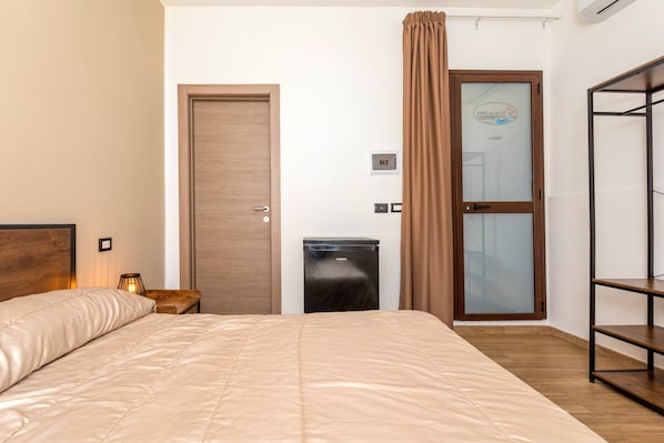 A bedroom with a bed, a wardrobe, a small desk with a lamp, a mini fridge, and a sliding glass door.