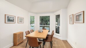 A dining area with a wooden table and brown chairs, framed pictures on the walls, and large windows with a view of trees.