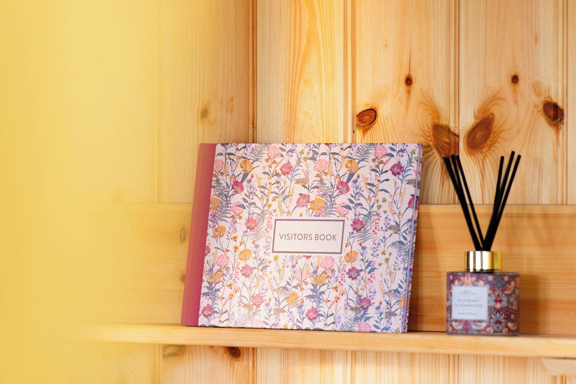 Un livre d’or est posé sur une étagère en bois ornée d’un motif floral, à côté d’un petit récipient contenant des diffuseurs à bâtonnets.