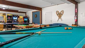 A pool table with billiard balls and cues, a dartboard, and arcade games in the background.