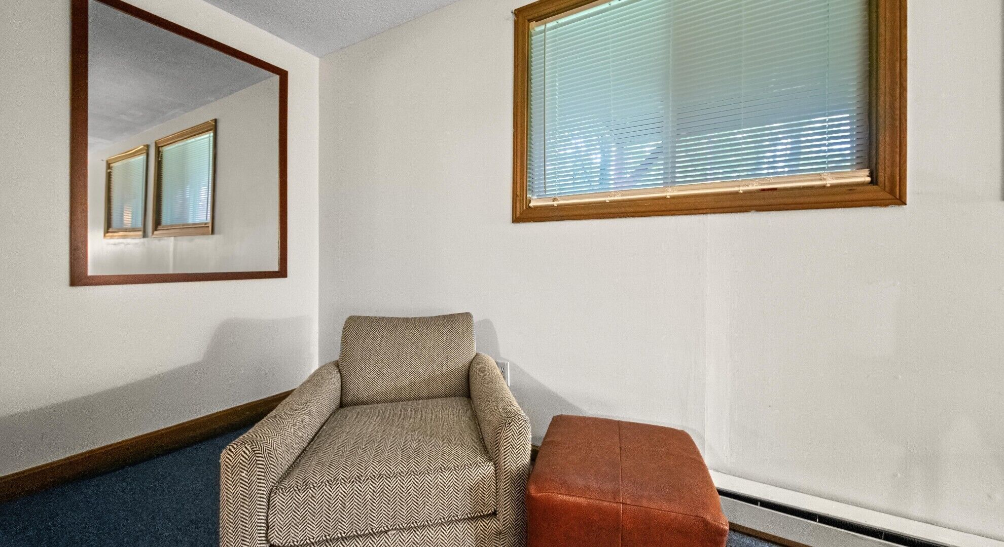 A room with a herringbone-patterned sofa, a small ottoman, a mirror, and a window with blinds.