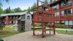 Une aire de jeux avec des structures d’escalade et une table de pique-nique devant un bâtiment doté de nombreuses fenêtres et d’une façade rouge.