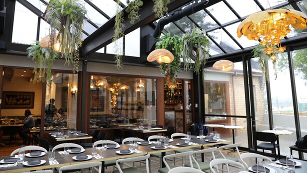 En restaurant med dækketøjl, hængende planter og et barområde, der kan ses gennem glasset.