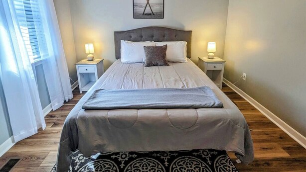 A bedroom with a bed, two bedside tables, a nightstand, a window with blinds, and a wall-mounted picture.