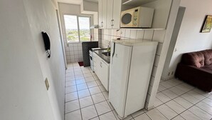 A compact kitchen with white cabinets, a microwave, a sink, and a stove. There is a brown couch in the adjacent living area. - Aquarius Flat Service (Caldas Novas)