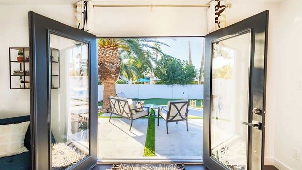 An open glass door leading to a patio with a table and chairs, a palm tree, and a view of a pool and greenery.