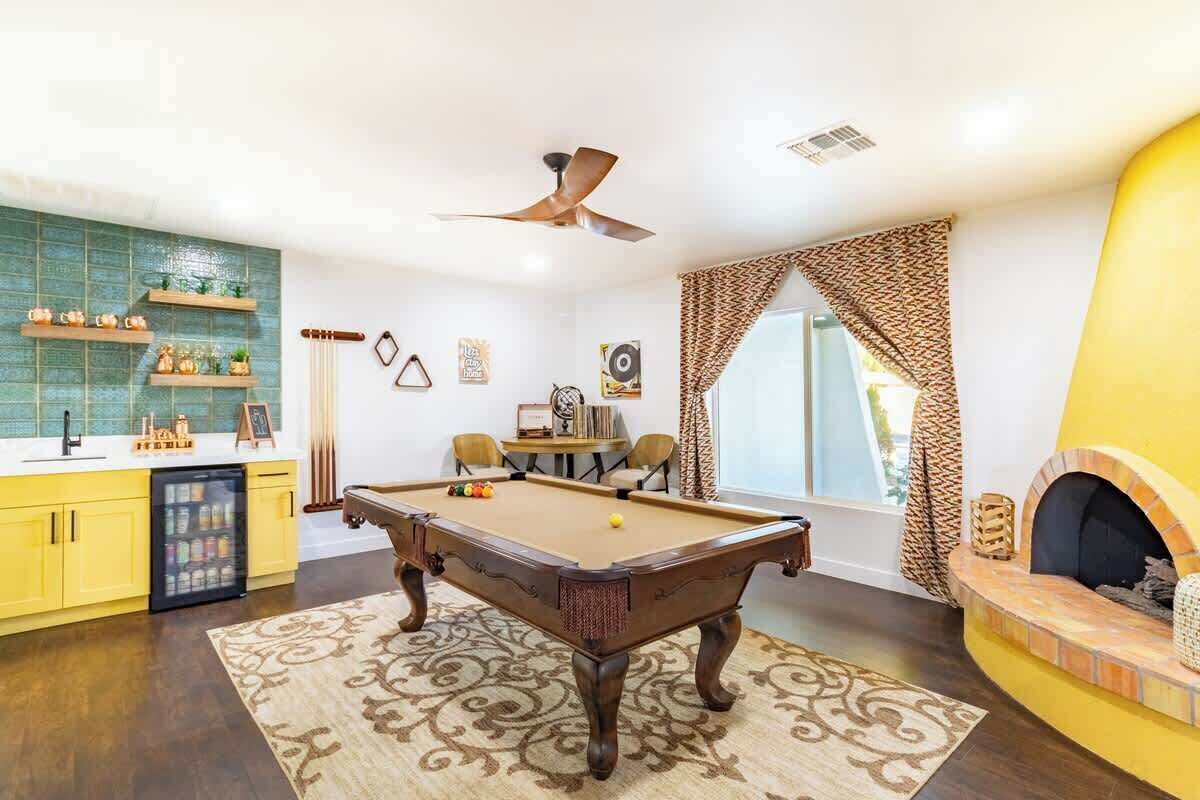 A pool table in a room with a yellow cabinet, a fireplace, and a window with patterned curtains.