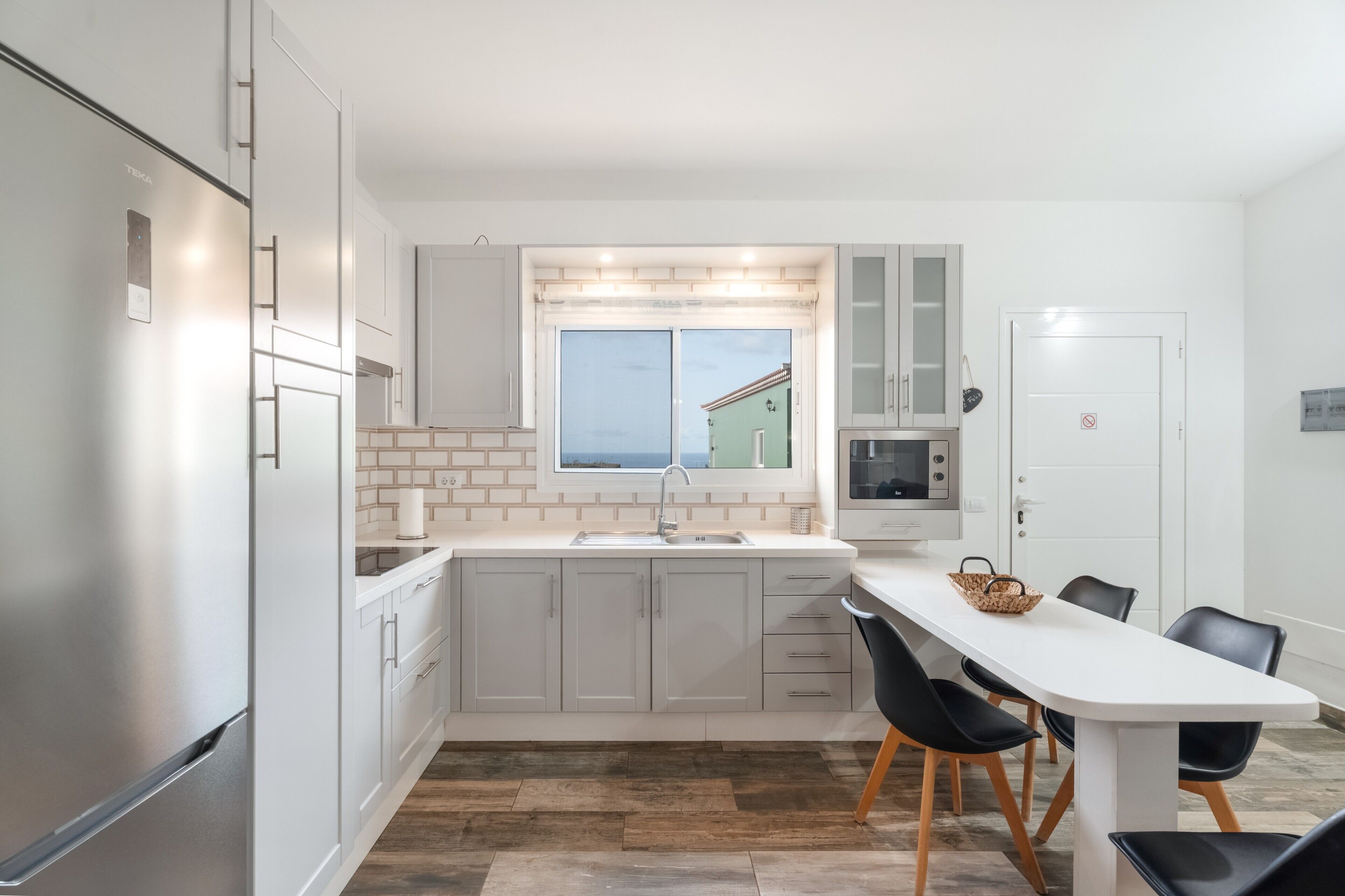 A modern kitchen with white cabinets, a stainless steel refrigerator, a window with a view, and a dining area with chairs.