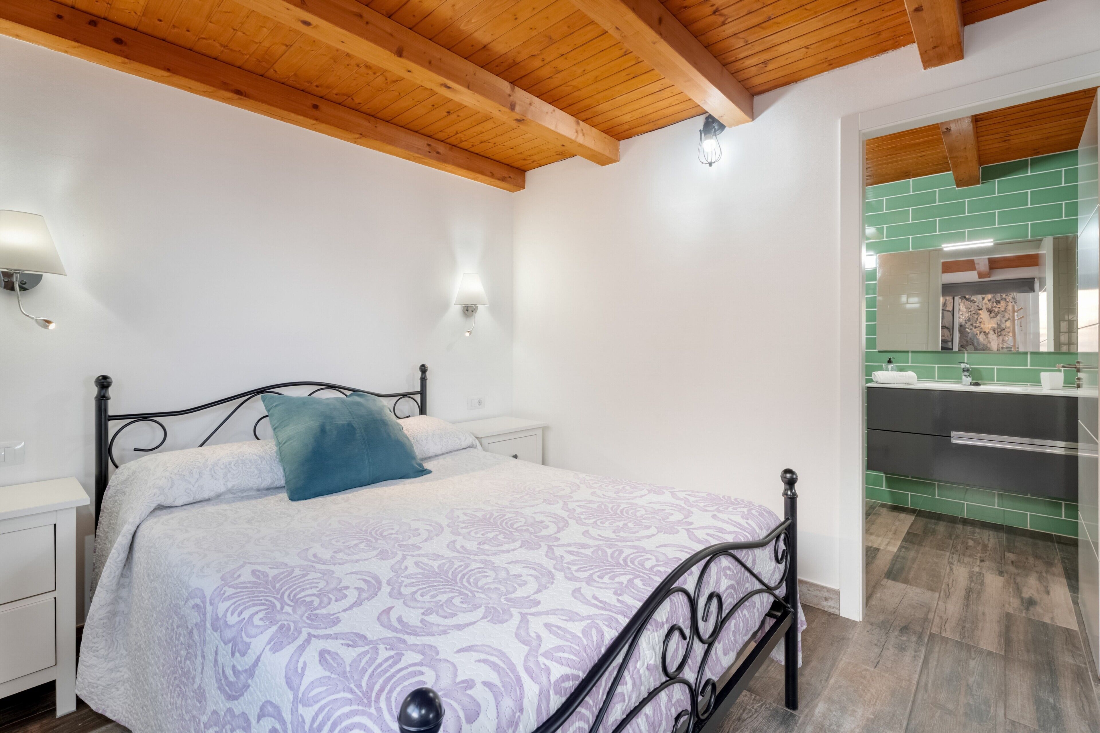 A bedroom with a bed, bedside tables, a nightstand, a green-tiled bathroom, and a wooden ceiling.