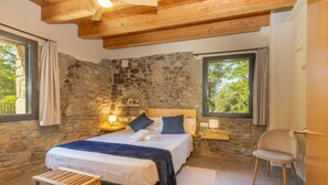 Une chambre avec un mur en pierre, des poutres en bois au plafond, un lit avec des oreillers, une table de chevet, une chaise et une fenêtre avec des rideaux.