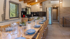 Un espace repas avec une longue table en bois dressée pour le repas, une cuisine équipée d’appareils modernes et un mur en pierre.