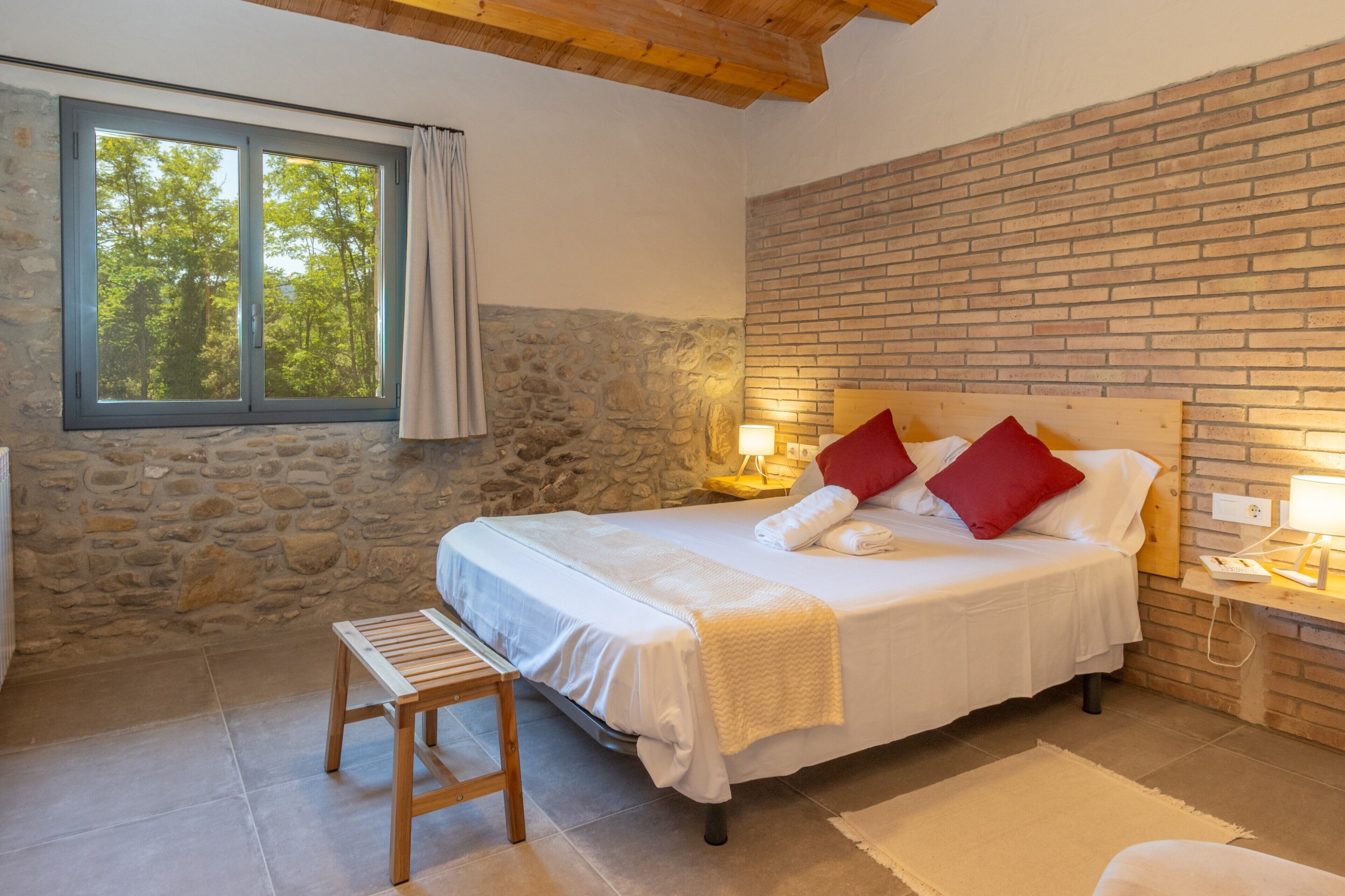 A bedroom with a stone wall, a wooden ceiling, a large bed with white bedding and red pillows, a bedside table with a lamp, and a window with curtains.