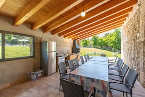 An outdoor kitchen with a stone wall, wooden beams, a long table, and wicker chairs. - Rural House 'La Rossa' with Mountain Views, Private Pool and Wi-Fi (Girona)