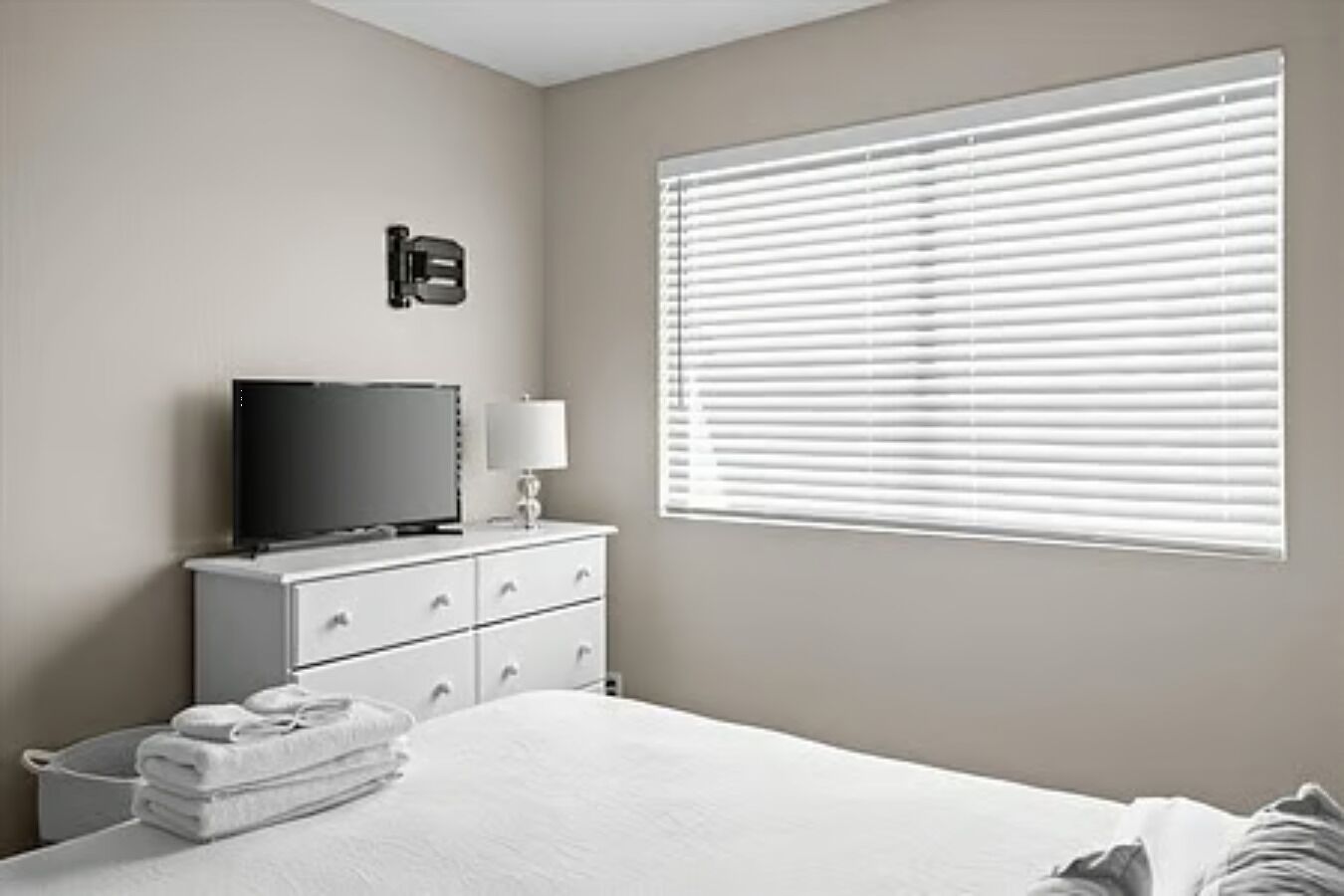 Een slaapkamer met een bed, een tv op een dressoir, een raam met jaloezieën en een wandlamp.
