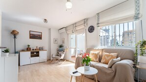 Ein modernes Wohnzimmer mit einer Couch, einem kleinen Tisch, einem Schaukelstuhl und einem Schrank. Ein Fenster bietet Blick auf Gebäude.