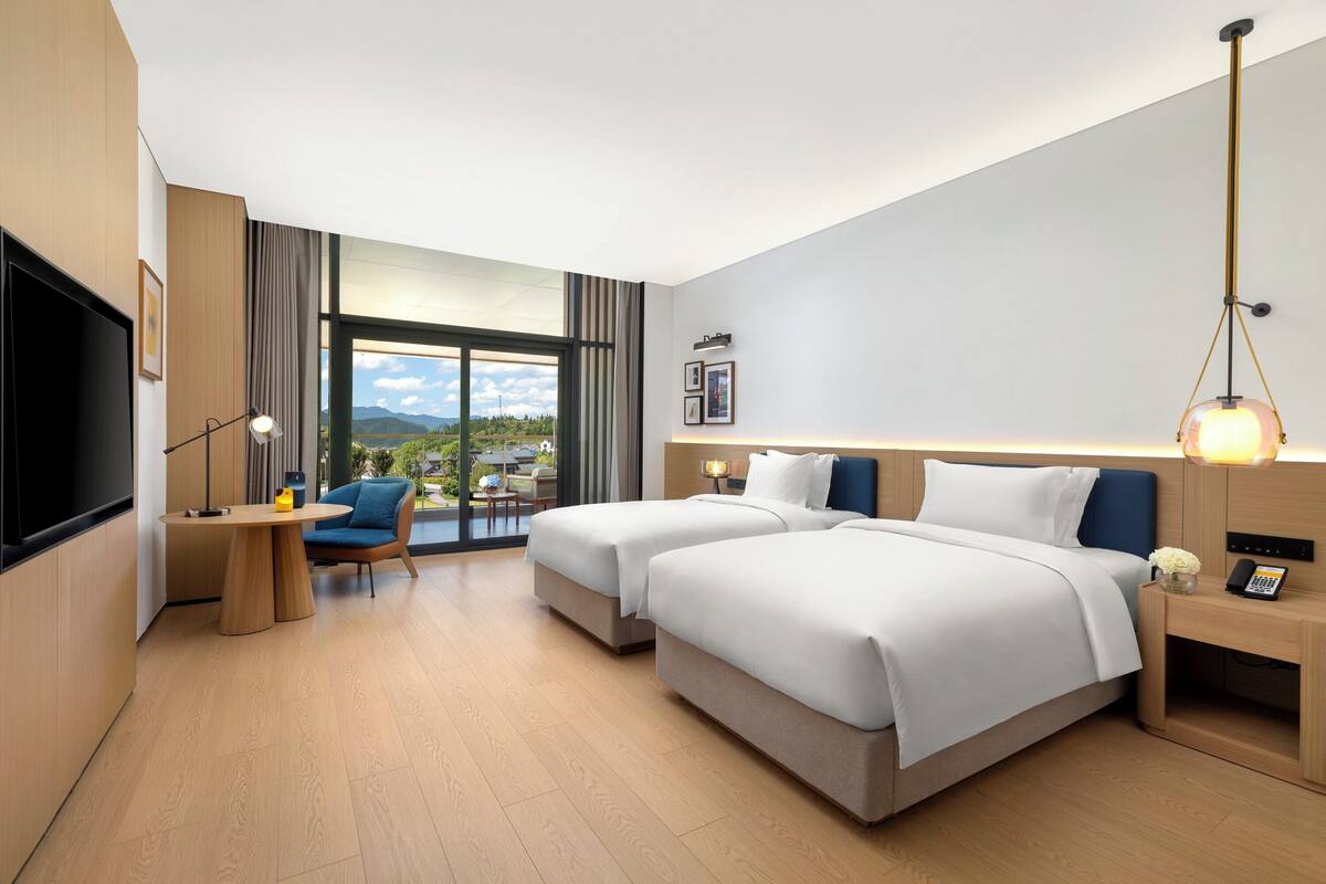 Une chambre d’hôtel moderne équipée de deux lits, d’un bureau, d’une chaise, d’une télévision et d’une grande fenêtre donnant sur l’extérieur.