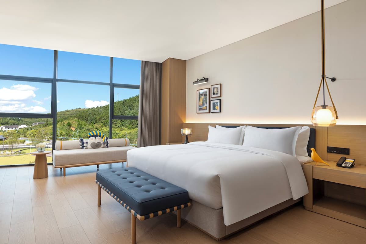 Une chambre d’hôtel moderne dotée d’un grand lit, d’une banquette, d’une table de chevet et offrant une vue sur un paysage verdoyant.