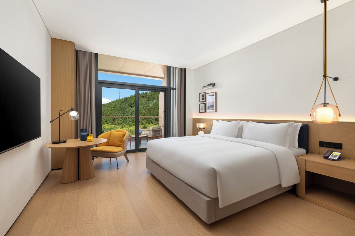 Ein modernes Hotelzimmer mit einem großen Bett, einem Schreibtisch mit Lampe, einem Sessel und Blick auf Grünflächen durch das Fenster.