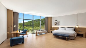 A modern hotel room with a large window, a sofa, a bed, a desk, and a bench.