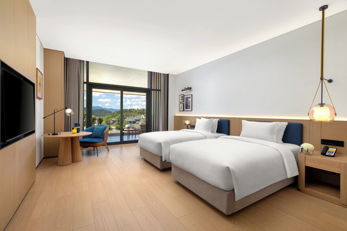 Ein modernes Hotelzimmer mit zwei Betten, einem Schreibtisch, einem Sessel, einem Fernseher und einem großen Fenster mit Blick ins Freie.