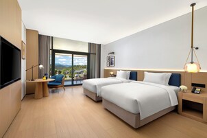Habitación de hotel moderna con dos camas, un escritorio, una silla, un televisor y un ventanal con vistas al exterior.