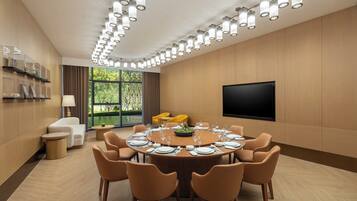 A modern conference room with a round table set for dining, surrounded by chairs, a sofa, and a TV on the wall.