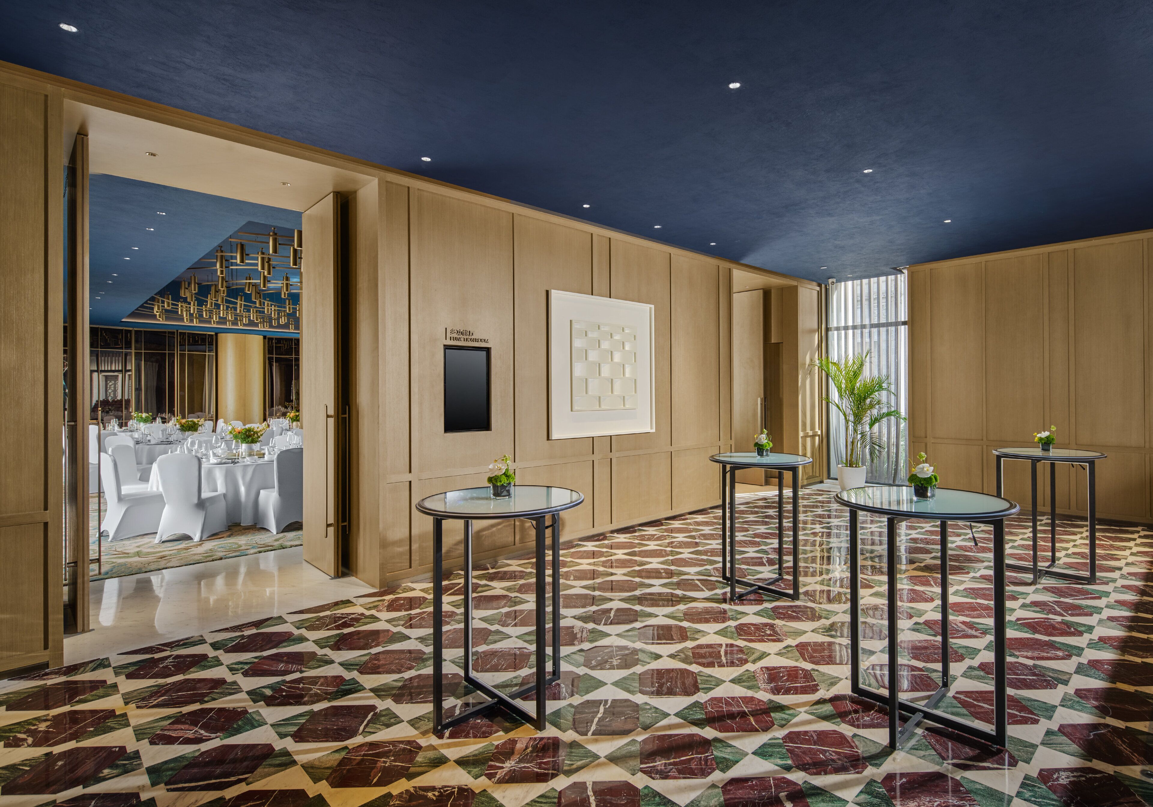 A modern hotel lobby with a geometric patterned floor, round tables with small plants, and a view into a dining area with white tables and chairs.