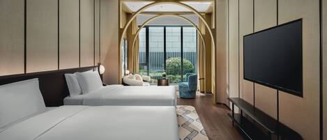 A hotel room with two beds, a TV, and a view of greenery through large windows.