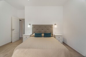 Une chambre à coucher avec une tête de lit capitonnée, deux lampes de chevet, un lit recouvert d’une couverture beige et une porte donnant sur une autre pièce.