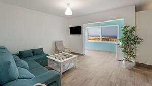 A modern living room with a sectional sofa, a coffee table, a chair, a TV mounted on the wall, a large window with a view, and a potted plant. - Holiday Home 'Elvira' with Sea View, Private Terrace and Wi-Fi (La Frontera)