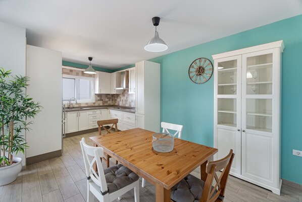 A dining area with a wooden table, white chairs, a potted plant, and a kitchen in the background.