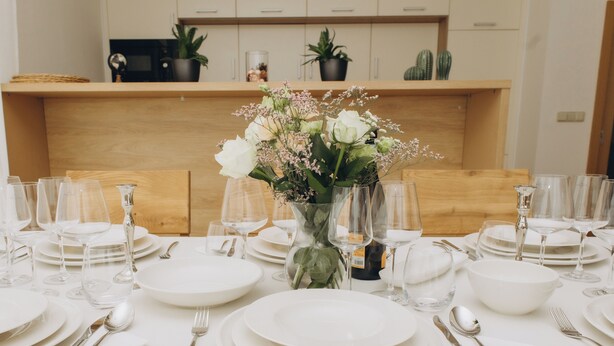 A dining table set with white plates, glasses, and silverware, centered with a vase of flowers.
