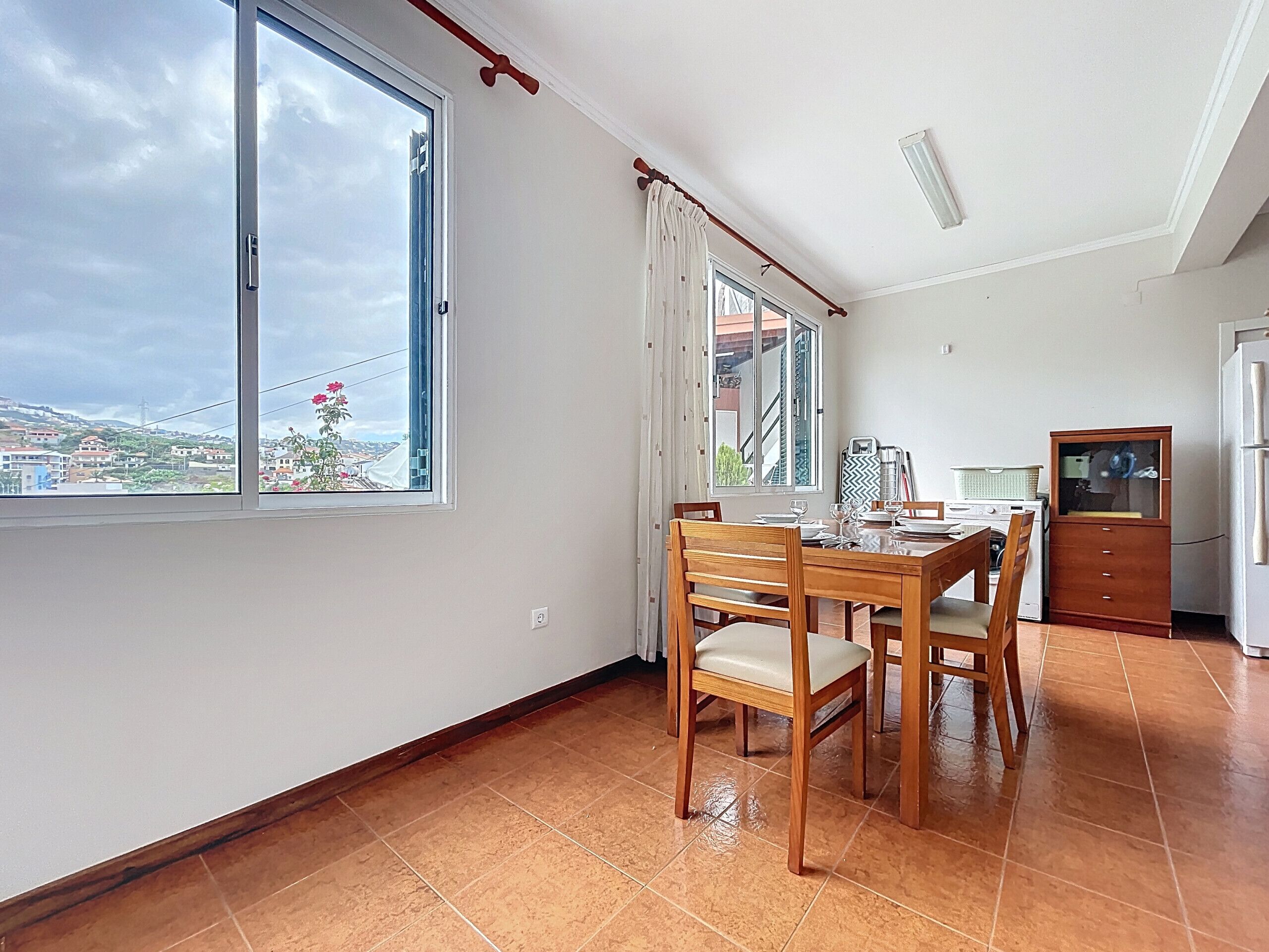 A dining area with a wooden table and chairs, a kitchen area with a refrigerator and stove, and large windows offering a view of the cityscape.