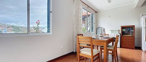 A dining area with a wooden table and chairs, a kitchen area with a refrigerator and stove, and large windows offering a view of the cityscape.