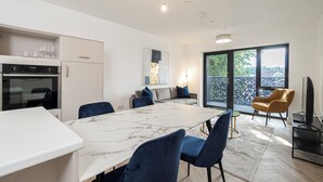 A modern kitchen with a marble countertop, a dining area with blue chairs, a sofa, and a TV. - Whispers in Light 65s Stay (Surrey)