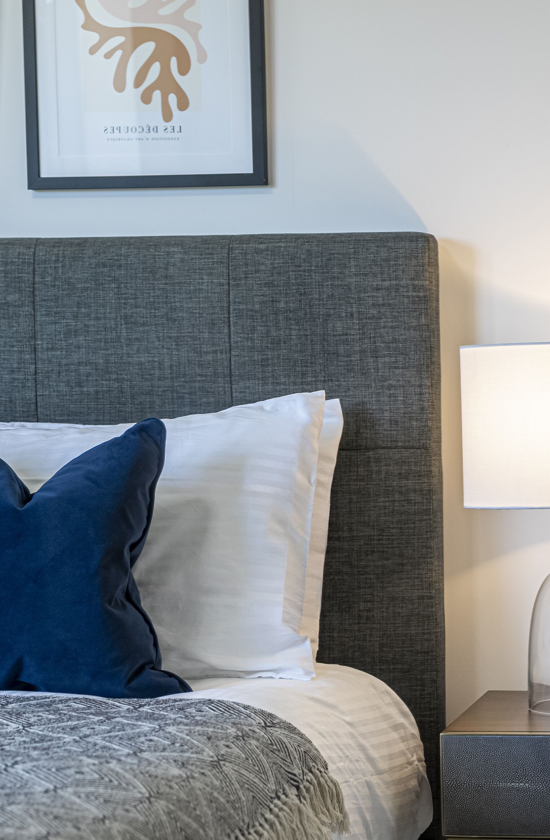 Un lit bien fait, avec une tête de lit grise, une lampe de chevet et une œuvre d’art encadrée accrochée au mur.